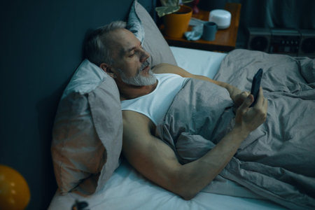 Senior man relaxing on bed while holding remote control and watching television. Cozy bedroom...