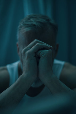 Close-up of a person holding head in hands while sitting in darkly lit room, showing emotions of...