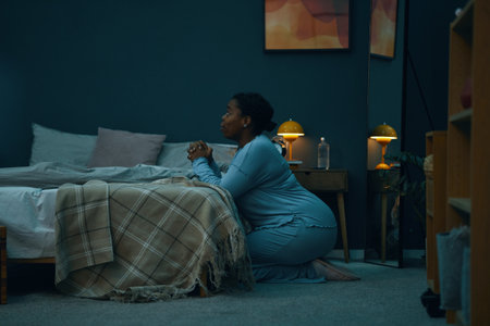 African American woman in blue nightgown praying next to bed in a dimly lit bedroom with artwork on walls and lamps illuminating sceneの写真素材