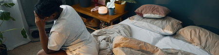 Person is sitting on edge of bed in bedroom, covering face with hand and other arm on knee,...