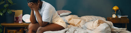 Woman sitting on bed with head in hands, seemingly overwhelmed, in cozy bedroom setting with nightstand and lamp in backgroundの写真素材