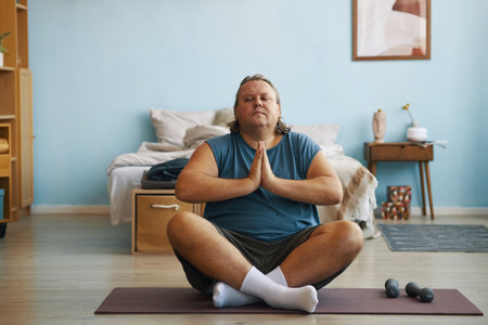 A man with closed eyes sitting cross-legged on yoga mat in his bedroom, hands in prayer position and focusing on inner peaceの写真素材