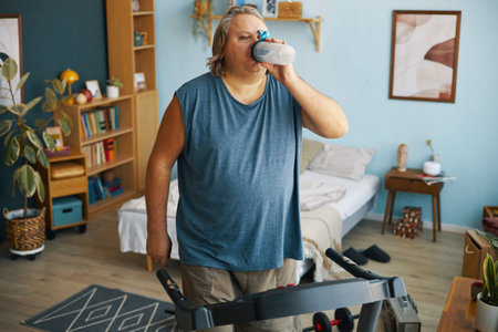 Man in casual clothing drinking water while exercising on treadmill in cozy, well-decorated living room with bookshelvesの写真素材