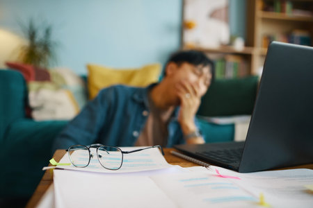 Blurred man sitting at wooden desk with head in hand, surrounded by papers and a laptop, creating an...