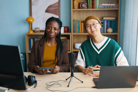 Young African American woman and Asian man sitting at desk with gadgets, smiling and collaborating...