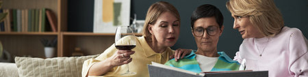 Three middle aged Caucasian women sitting together on sofa, one holding wine glass, all looking at...
