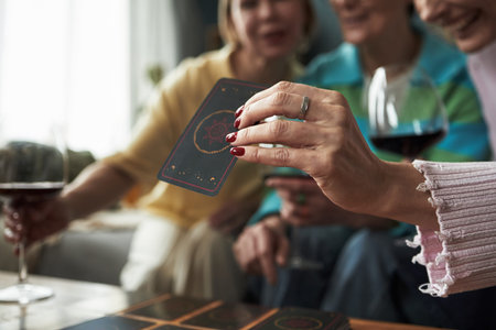 Group of young adult Caucasian women playing card game together, holding cards and drinking red wine, close up of hand with ring in foreground, friends enjoying leisure activity indoorsの写真素材