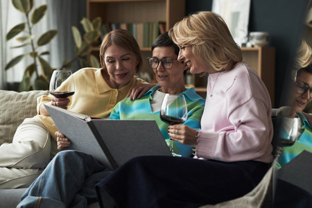 Three Caucasian middle aged women sitting together on sofa smiling and looking at large photo album while holding wine glasses, enjoying friendly conversation and bondingの写真素材