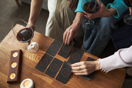 Three young adult Caucasian women sitting together holding glasses of red wine, playing tarot card game on wooden table, hands reaching for cards, candles burning nearbyの写真素材