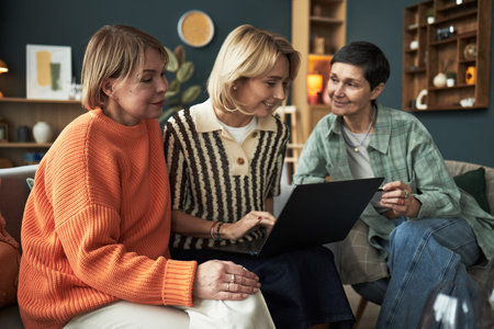 Three middle aged Caucasian women sitting together on sofa using laptop, smiling and interacting,...