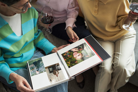 Three middle aged Caucasian women sitting together holding glasses of red wine, looking at photo...