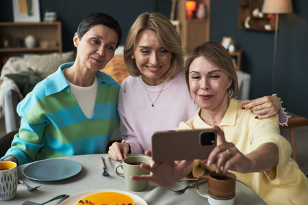 Three Caucasian middle aged women sitting together at table smiling and taking selfie with...