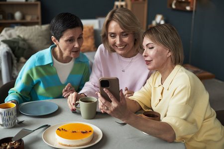 Three middle aged Caucasian women sitting at table smiling and looking at smartphone together,...