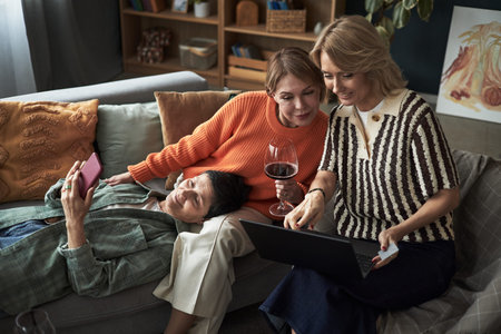 Three Caucasian young adult women relaxing on sofa, one lying down holding smartphone, two sitting...