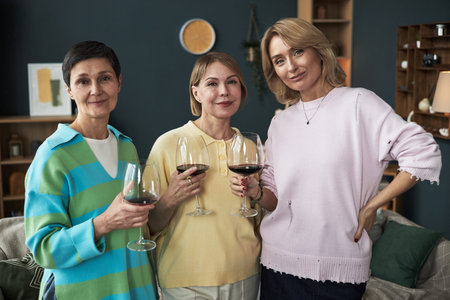 Three middle aged Caucasian women standing together holding wine glasses, smiling at camera, showing friendship and connection in casual indoor setting, celebrating adult girlfriendsの写真素材
