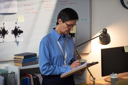 Middle aged Caucasian woman with short dark hair standing in office holding clipboard, writing notes...