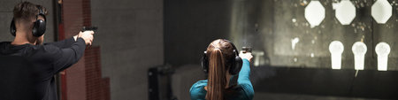 Young Caucasian man and woman standing side by side aiming handguns at paper targets in indoor...
