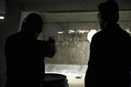Dark silhouettes of two adult men standing at indoor shooting range, aiming handgun at target and observing, both wearing protective ear muffs and safety glassesの写真素材