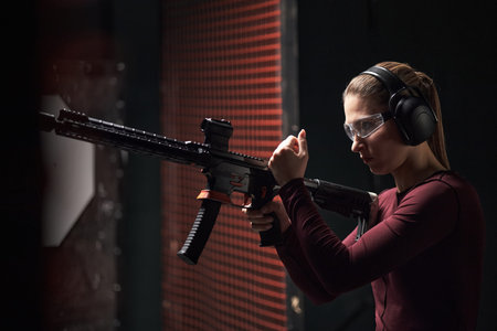 Caucasian young woman wearing protective glasses and earmuffs, holding rifle, preparing to shoot at...