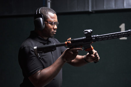 Black man young adult wearing protective earmuffs and safety glasses aiming modern rifle at indoor...