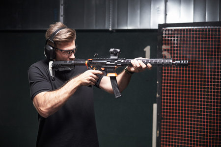Caucasian male customer wearing protective earmuffs aiming modern rifle at indoor shooting range,...
