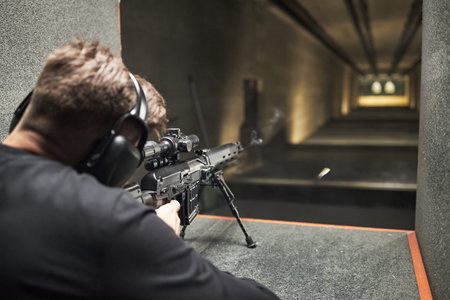 Rear view of man wearing hearing protection aiming rifle at targets in indoor gun range, bullet...