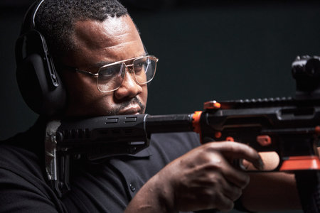 Black middle aged man wearing glasses and earmuffs aiming rifle at indoor shooting range, focusing...
