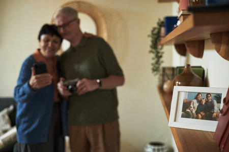 Senior couple standing closely, smiling, and posing with smartphone in cozy living room. Background includes framed photo on shelf and warm home decor creating a welcoming environmentの写真素材