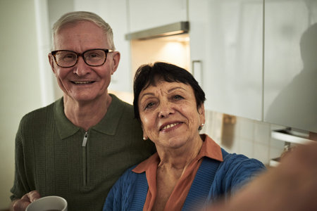 Portrait of elderly couple smiling while posing for selfie in bright kitchen. Man wearing glasses...