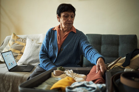 Elderly woman packing clothes in suitcase while preparing to go on vacation, sitting on couch with laptop open in backgroundの写真素材