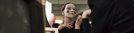 Caucasian teenager girl applying face makeup and red blush with brush while sitting at home,...
