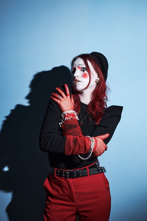Caucasian teenager girl with red hair and artistic makeup standing against blue wall, crossing arms...