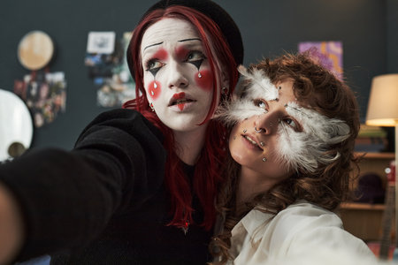 Two creative Gen Z girls, one Caucasian teenager with dramatic clown makeup and red hair, one Caucasian teenager with feather face art and piercings, taking selfie together at homeの写真素材