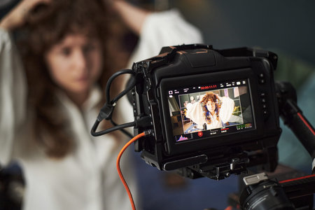 Caucasian young adult woman styling hair in front of professional video camera, camera screen...