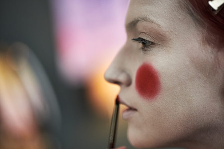 Caucasian young adult woman applying bold red makeup to face using brush, focusing on cheek and...