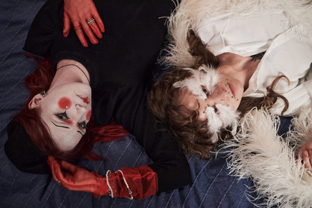 Two Caucasian teenage girls lying on bed, wearing creative makeup and accessories, one with dramatic...
