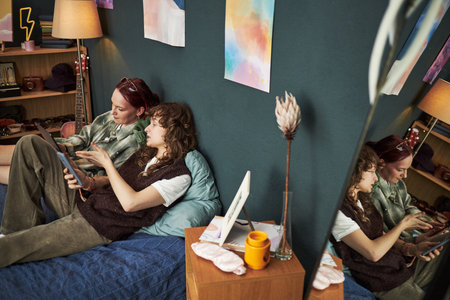 Two Caucasian teenage girls sitting on bed using digital tablet together, one girl leaning against wall, both focused on screen, creative posters and art supplies visible in backgroundの写真素材