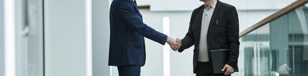 Header of businessmen shaking hands in a modern office with glass walls and staircase. Professional attire, indicating a successful meeting, is visible in the interactionの写真素材