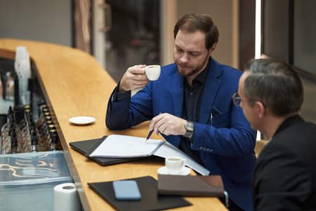 Two businessmen reviewing documents and having coffee at office counter, engaging in professional...
