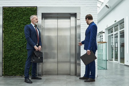 Two businessmen standing near elevator in a modern office building. One man is holding briefcase...