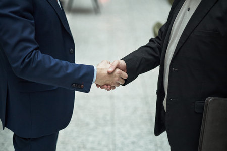 Businessmen in formal attire shaking hands while standing in office lobby, focusing on agreement and...