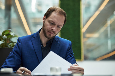 Serious young male manager in blue blazer closely reviewing document at modern workplace with green...