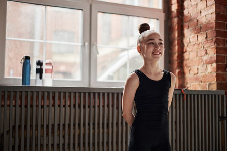 Portrait of Caucasian girl child smiling during rhythmic gymnastics training standing in gym with hands behind back, hair in bun, natural light coming through large window in backgroundの写真素材