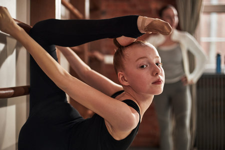 Portrait of Caucasian teenage girl practicing advanced flexibility pose during rhythmic gymnastics training in studio, stretching leg over head while instructor observing in backgroundの写真素材
