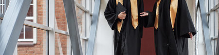 Header of two anonymous young students wearing graduation gowns standing in hallway talking and holding diploma folders, university students engaging in conversation after ceremonyの写真素材