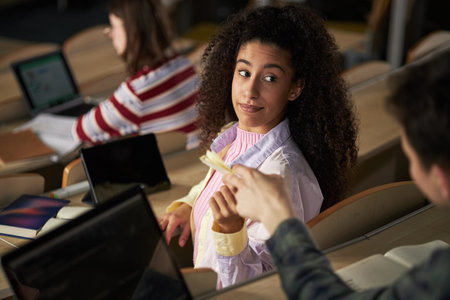 Young Black woman sitting in university lecture hall turning to look at Caucasian group mate...