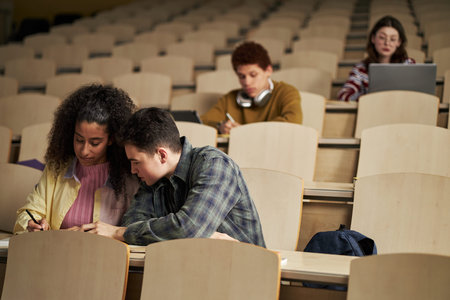 Multiethnic group of young university students sitting in lecture hall, two students collaborating...