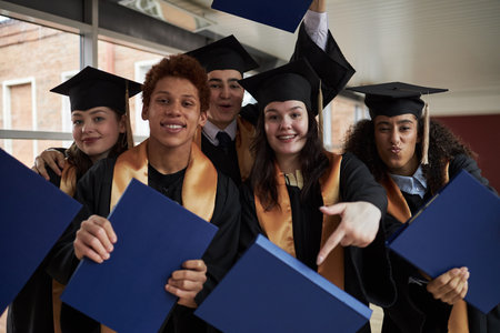 Group of multiethnic teenagers wearing graduation caps and gowns celebrating university achievement, holding diploma folders, smiling and posing together for camera indoorsの写真素材