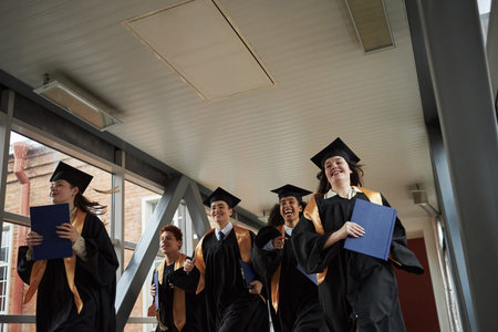 Group of multiethnic young university students wearing graduation caps and gowns running down...