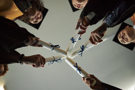 Group of diverse young university students wearing graduation caps holding diplomas together in circle celebrating academic achievement viewed from belowの写真素材